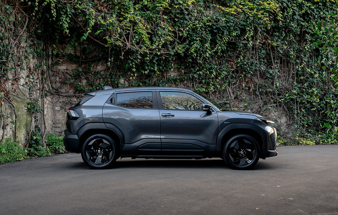 A dark grey compact SUV taxi is parked on a paved road, with a wall covered in green ivy and plants in the background.