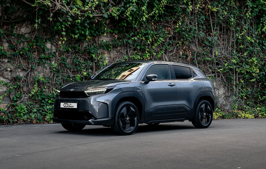 A sleek grey SUV taxi is parked on a paved road, with a stone wall covered in green ivy in the background.