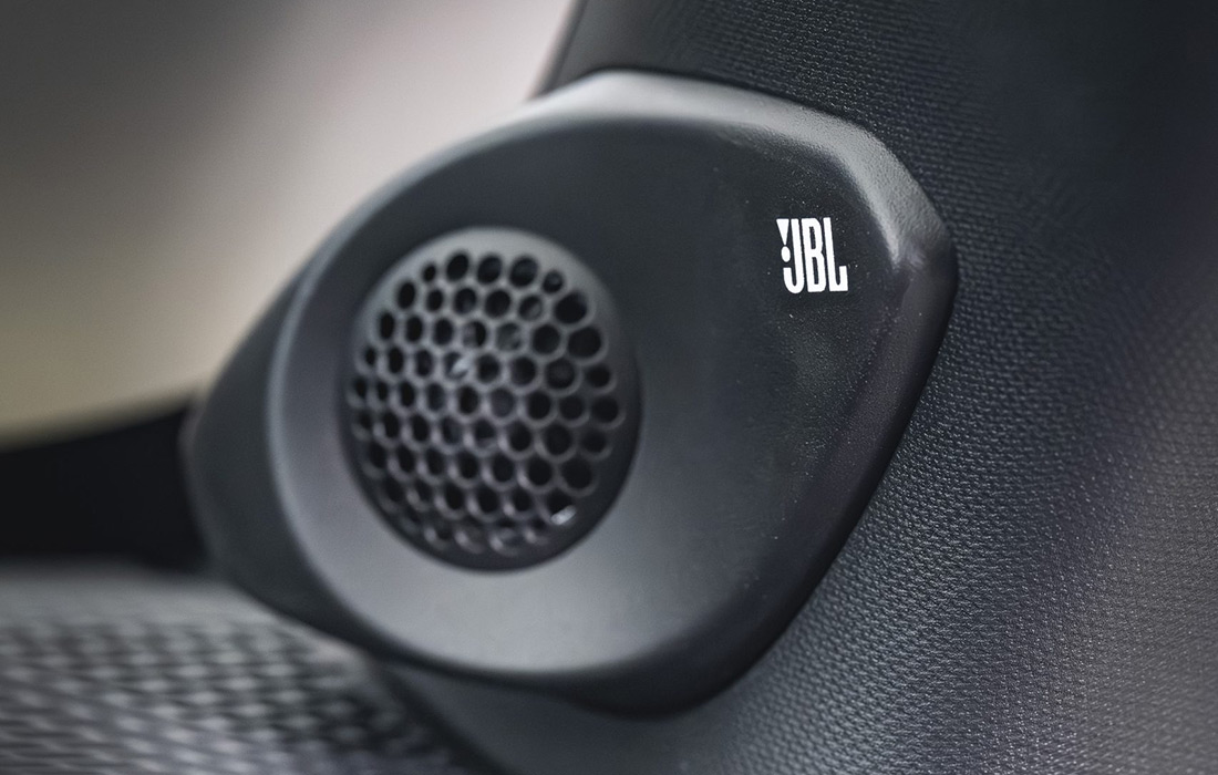 Close-up of a black JBL car speaker with a honeycomb grille, mounted on a textured black surface inside a taxi. The white JBL logo is visible on the speaker.