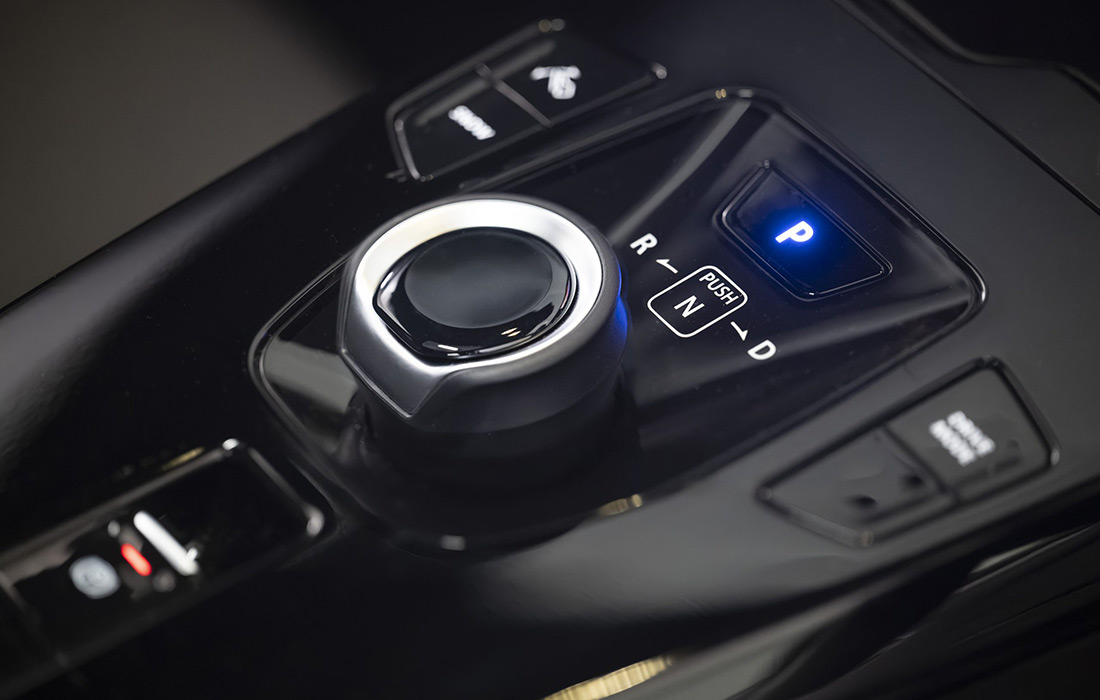 Close-up of a modern car gear selector with a rotary knob and illuminated P button for park. Other labelled positions include R, N, and D, with surrounding black glossy surfaces and adjacent control buttons.