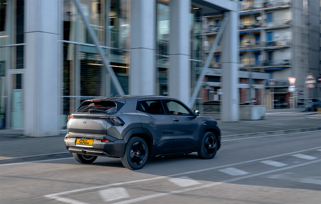 A grey compact SUV taxi drives on a city street past modern glass buildings and flats, rear end visible with slight motion blur.