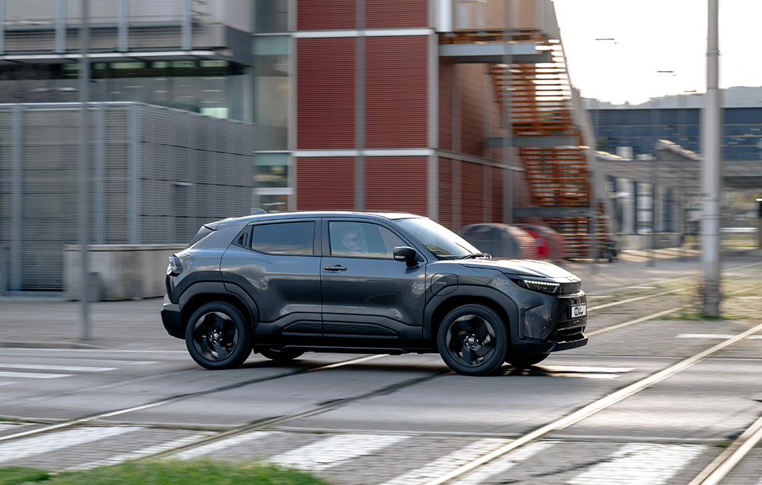 A dark grey compact SUV taxi drives past modern buildings, crossing tramlines on a city street in daylight.