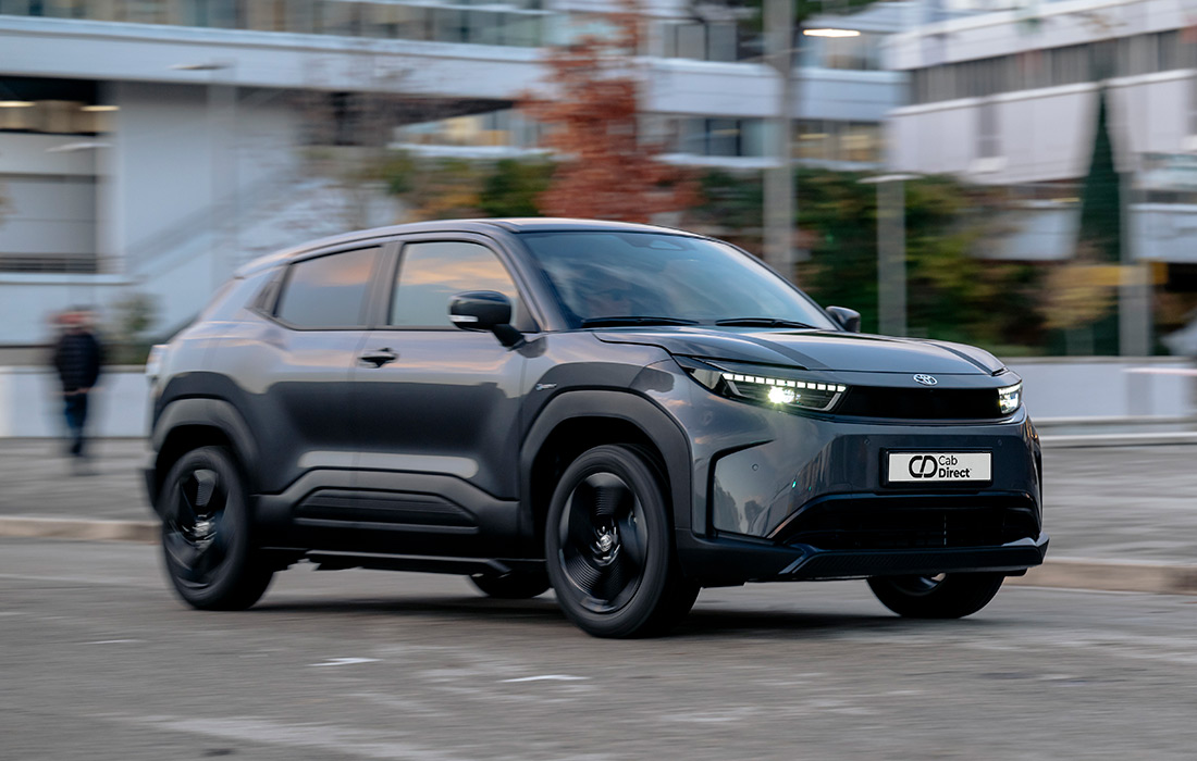 A sleek, dark grey compact SUV taxi drives through a city street with blurred buildings and trees in the background, highlighting motion. Featuring modern LED headlamps and a distinctive, futuristic design—ideal for urban taxi services.