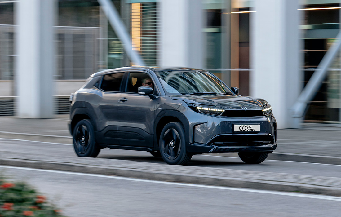 A sleek, dark grey SUV taxi drives through a modern city with motion-blurred buildings in the background.