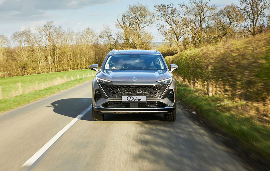 A silver SUV taxi drives down a rural road lined with green fields and trees under a partly cloudy sky, viewed from the front with a number plate that reads Car Direct.