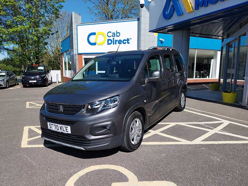 A grey taxi is parked in a disabled parking bay outside the Cab Direct dealership on a sunny day, with the dealership sign and building visible in the background.
