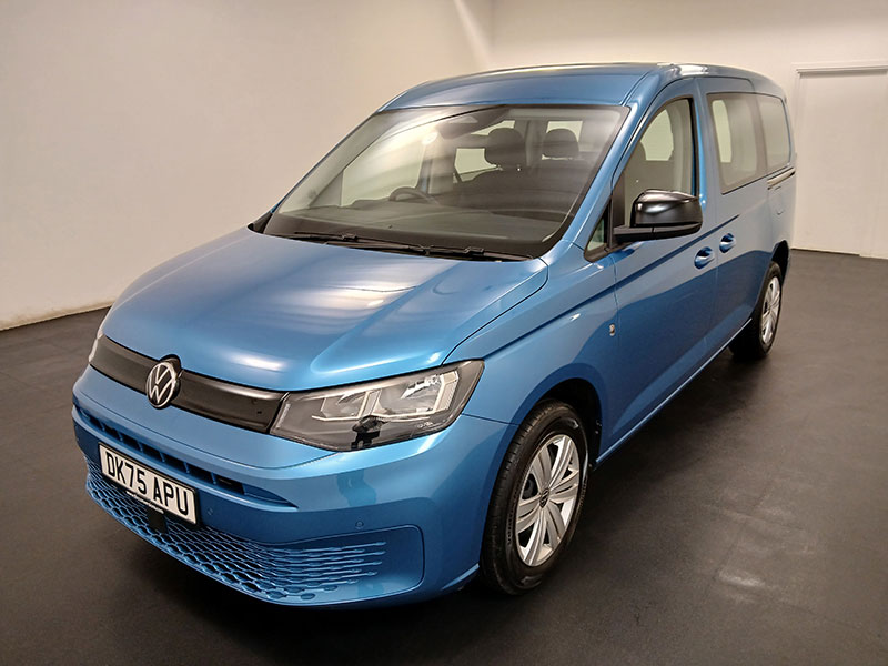 A blue Volkswagen taxi is parked indoors on a black floor against plain white walls, shown from the front left side highlighting its modern design and headlamps.