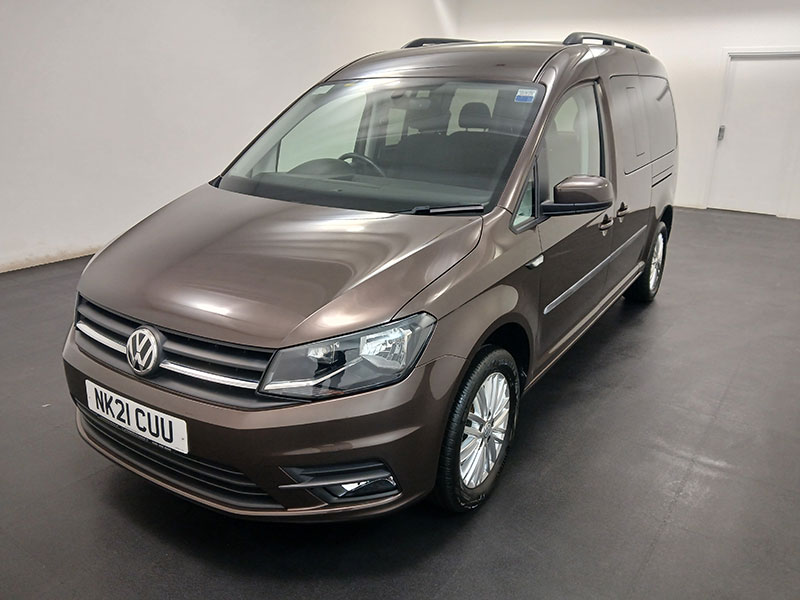 A brown Volkswagen taxi is parked indoors on a black floor, facing left. It features UK number plates and is shown under bright, even lighting.