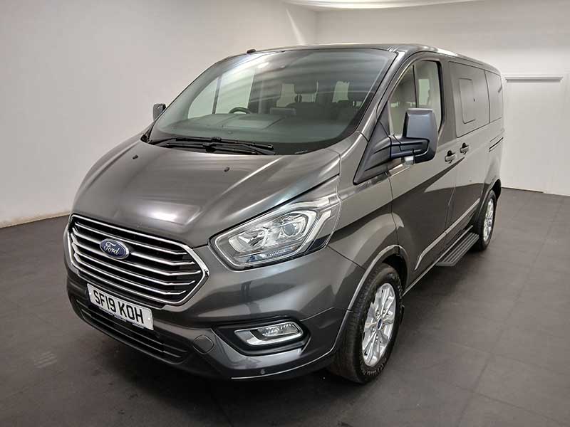 A grey Ford Transit taxi is parked in an indoor showroom with a plain white wall and black floor, shown from the front left side highlighting its headlamps and grille.