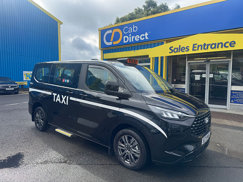 A black taxi is parked outside the Cab Direct Sales Entrance, with the building’s blue and yellow façade visible under an overcast sky.