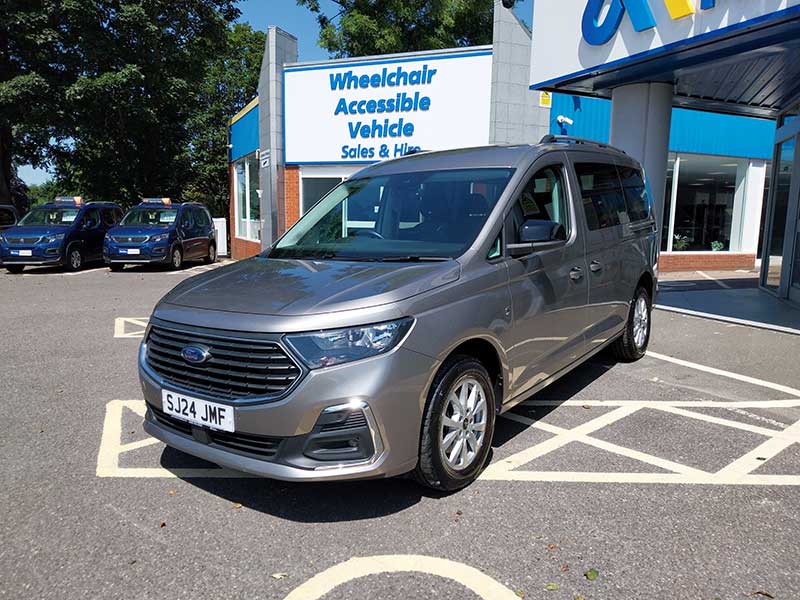 A silver wheelchair accessible taxi is parked in a designated accessible parking space outside a dealership labelled Wheelchair Accessible Vehicle Sales & Hire.