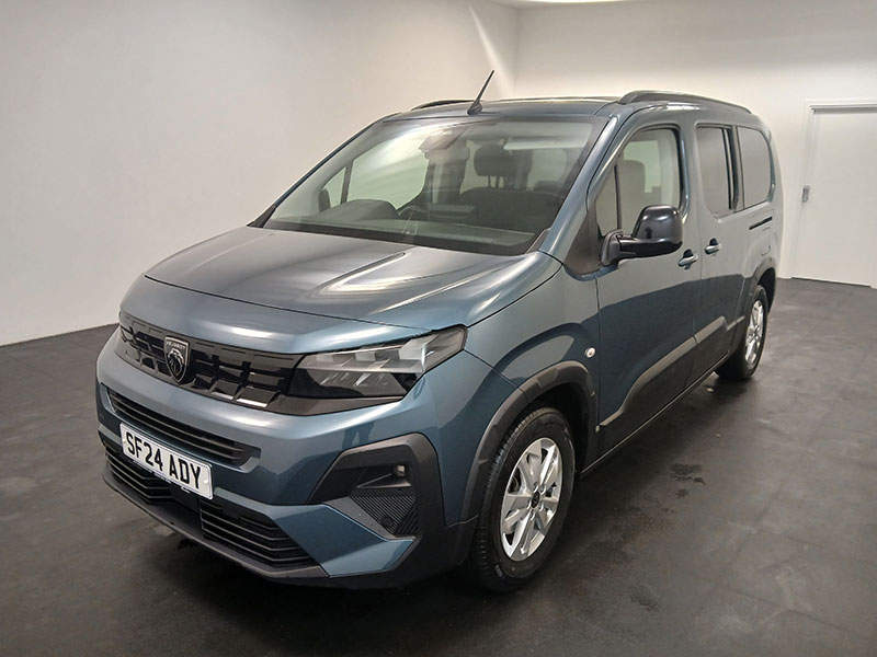 A blue Peugeot Rifter taxi is parked indoors on a dark floor, angled to the left. Its number plate reads SF24 ADY. The setting features plain white walls and a door in the background.