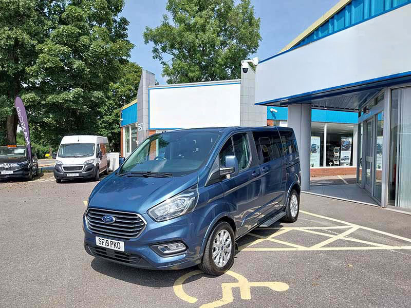 A blue Ford taxi is parked in a disabled parking bay outside a building with glass doors, with trees and other vehicles nearby on a sunny day.