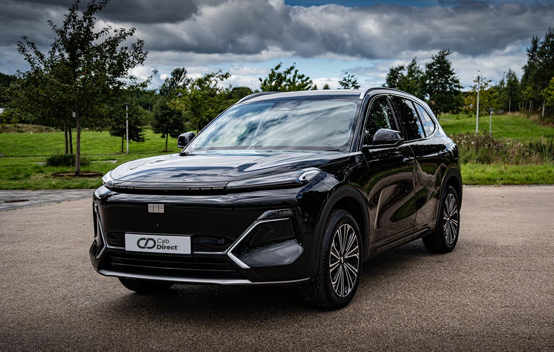A sleek black SUV taxi is parked on a paved area with green trees and grass in the background under a cloudy sky.
