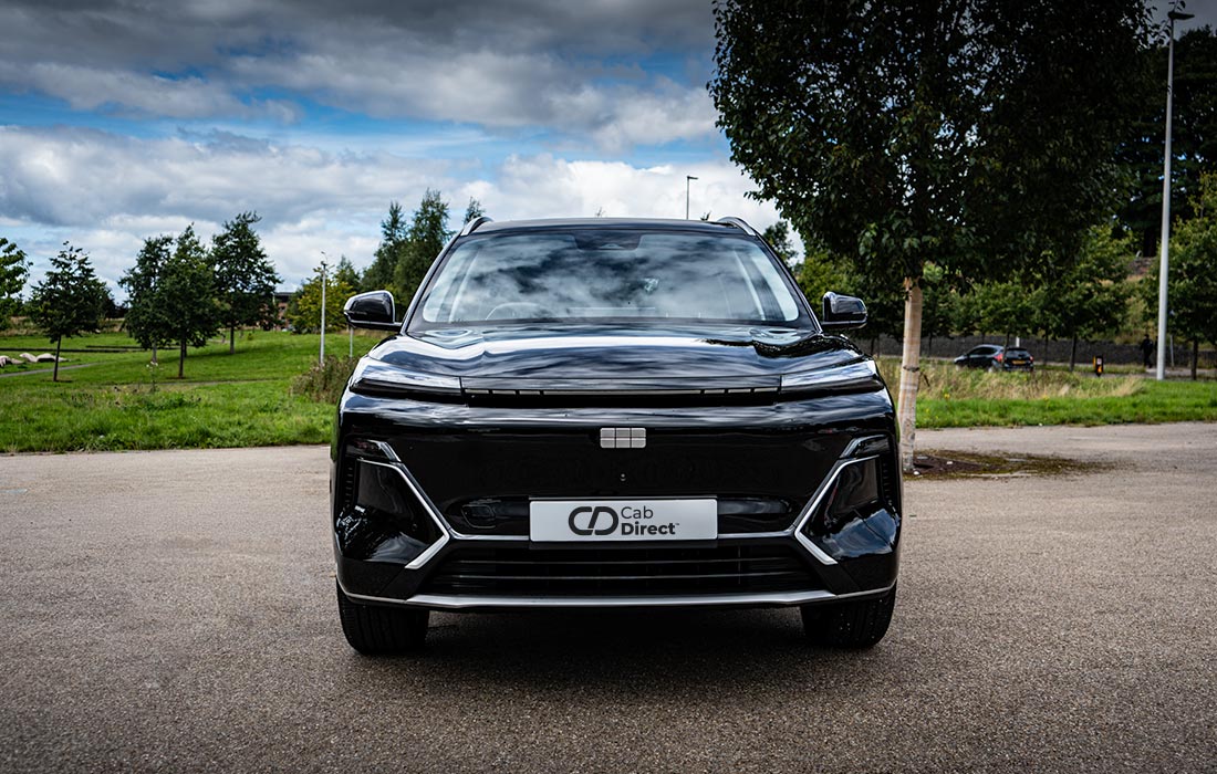 A black SUV taxi is parked on a paved area with green trees and grass in the background. The vehicle faces forwards, displaying a Cab Direct number plate under a partly cloudy sky.