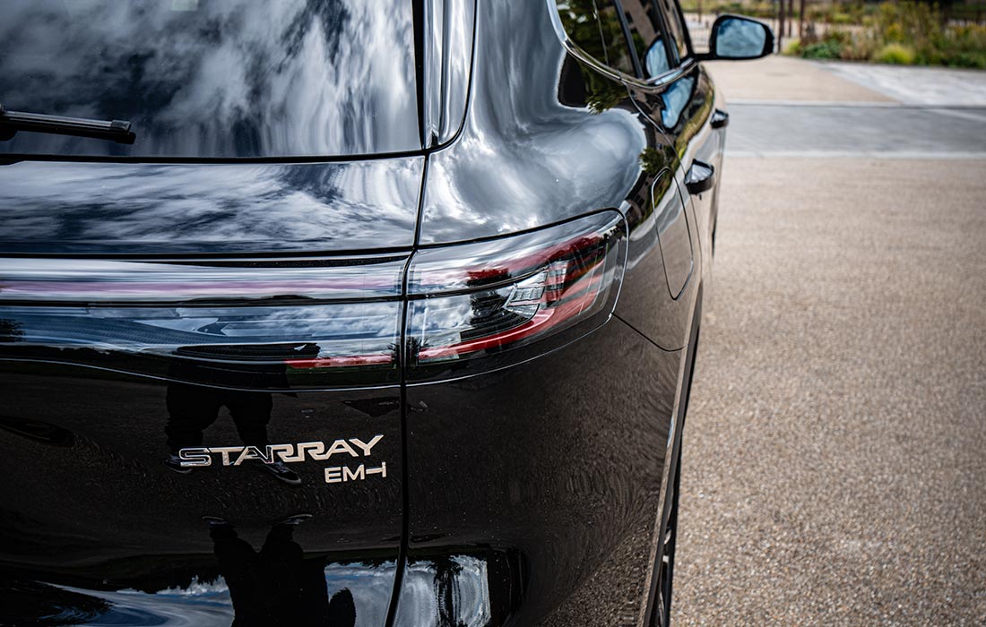 Rear view close-up of a sleek black STARRAY EM-H taxi, showcasing its distinctive badges and reflective finish, parked on paved ground with trees and sky mirrored in the car’s surface.
