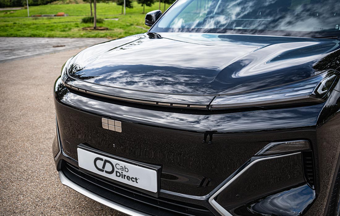 Close-up of a shiny black electric taxi front, highlighting sleek headlights, a smooth grille, and a Cab Direct number plate, parked outdoors on paved ground with grass in the background.