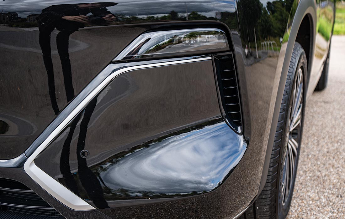 Close-up of a black taxi’s front bumper and headlamp, highlighting clean lines and reflective surfaces with visible sky, clouds, and the photographer in the reflection. Part of the front tyre and road are also shown.