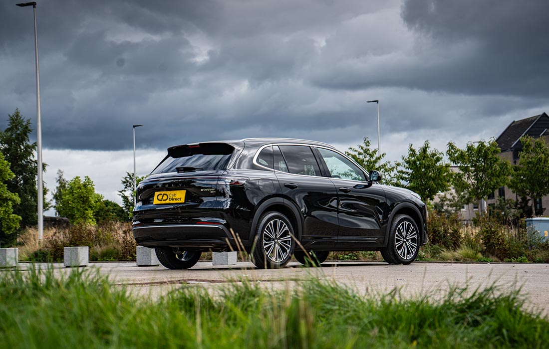 A black SUV taxi is parked on a paved car park surrounded by greenery. Overhead, dark clouds are visible, with trees and buildings in the distance.