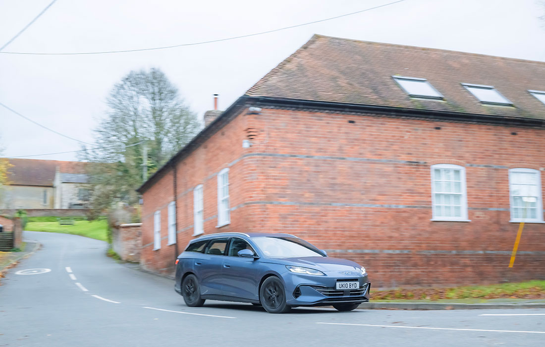 Blue MG5 estate taxi driving along a rural road past a brick building.