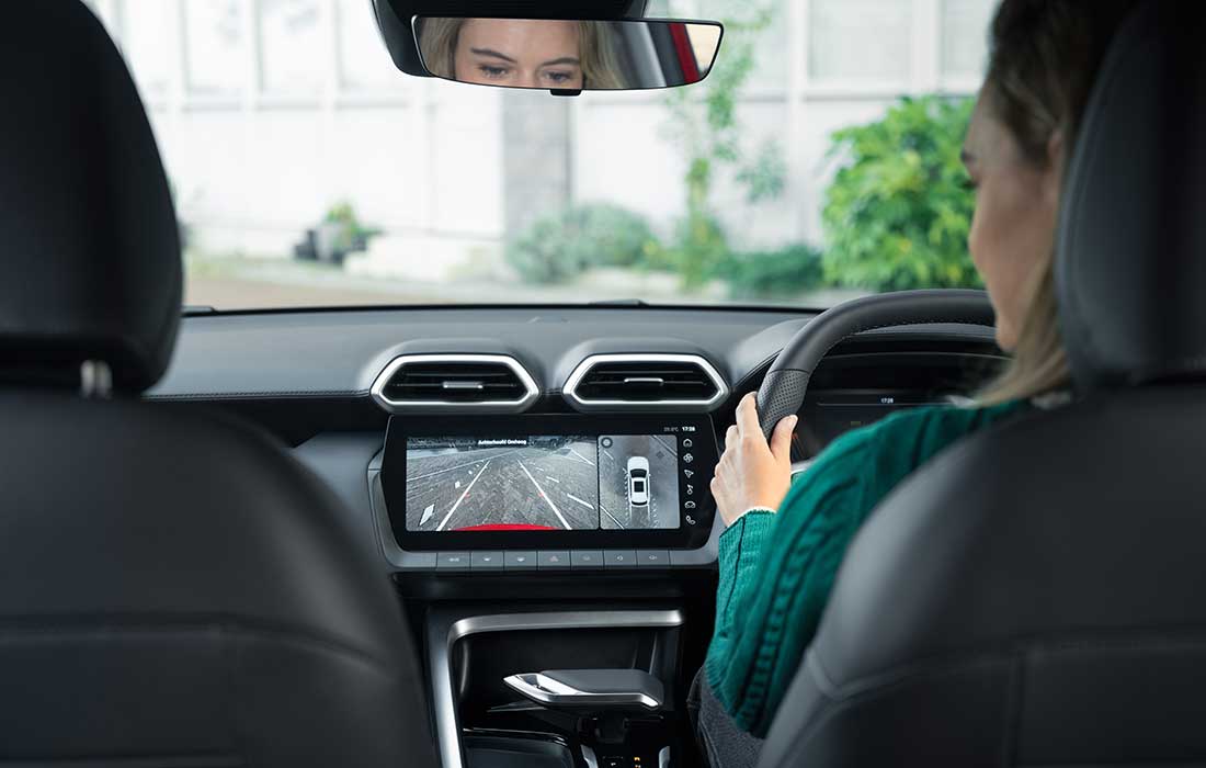 Interior view from the rear passenger area of a taxi, showing the driver looking at the centre dashboard screen displaying the rear-view camera.